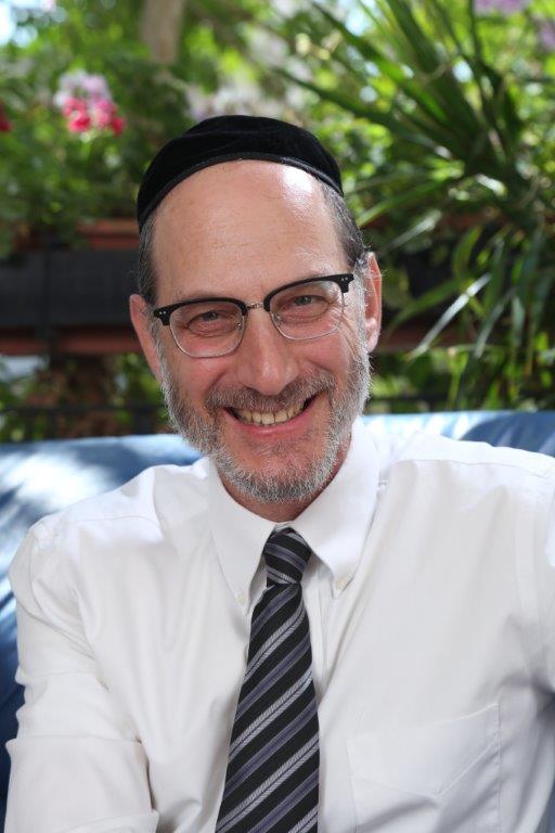 Smiling man wearing a black kippah, glasses, white shirt, and striped tie outdoors.